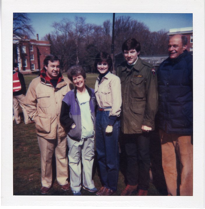 The image features five individuals -- two women and three men -- posing together on a grassy lawn on the Mary Washington campus. 