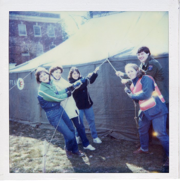 The image features five individuals -- three women and one man -- pulling on two ropes attached to a large tent. There are three people on the left rope and two on the right. The tent is located on a grassy  lawn on the Mary Washington campus. 