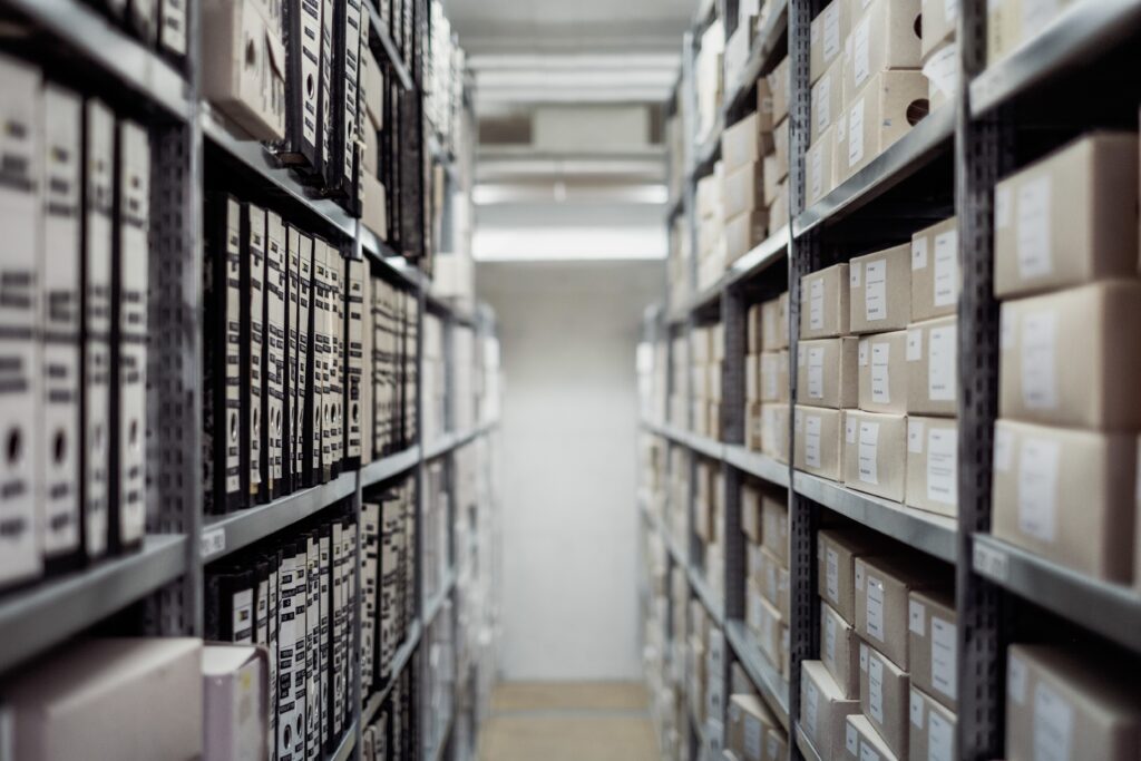 A close-up photograph of a shelf filled with white paper document binders.and boxes full of archival papers.