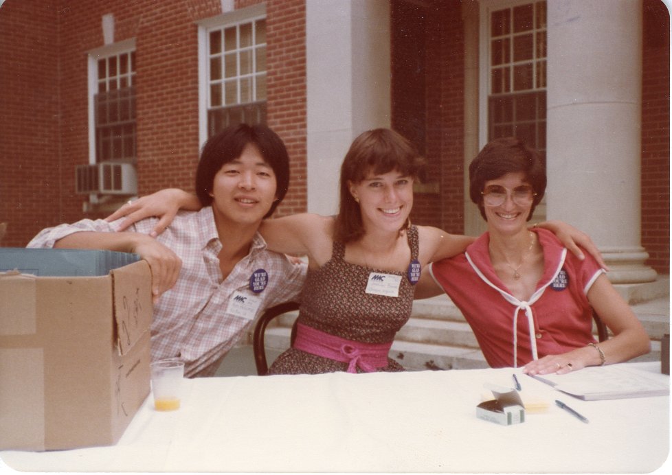 This image features three individuals -- one man and two women -- sitting together, posing for the picture, at a table with a white tablecloth. The people are all wearing blue pins with say "WE'RE GLAD YOU'RE HERE." The table has a cardboard box on it and a few pens scattered around. 