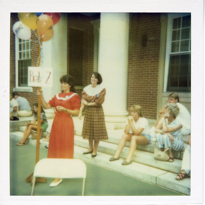 This photo features a group of people gathered on wide concrete steps at the base of a large brick building with white columns. Most of the people are looking at one girl in the front, in a red dress, who is holding a sign that says "Brah-Z."