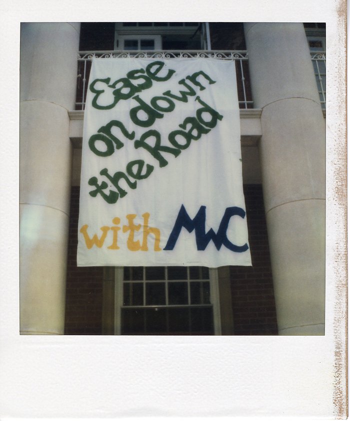 A sign hanging off of a white railing between two white columns. The sign reads "Ease on down the road with MWC." 