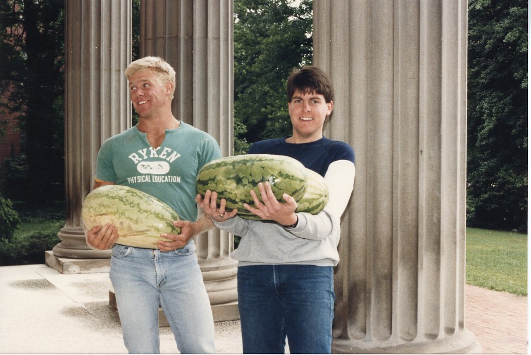 This photo features two men, who are carrying very large watermelons. They are posed in front of very large white columns. 