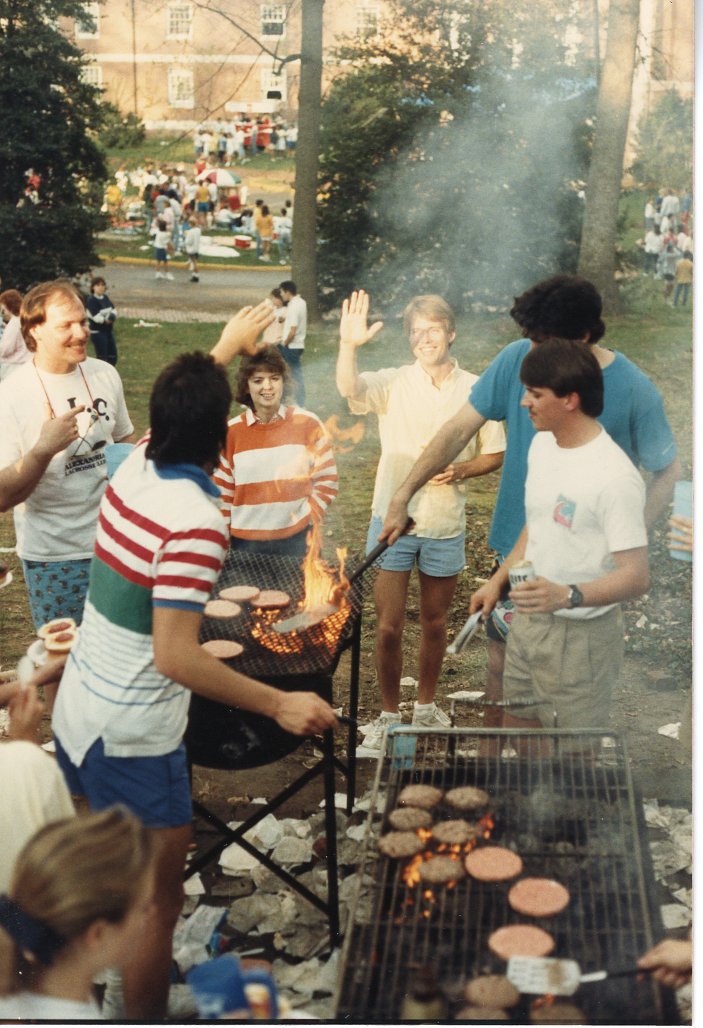 This photo depicts a group of people who are hanging out around two grills. A few of them are grilling, while the rest either talk to each other or eat. On the grills are hamburger patties. 