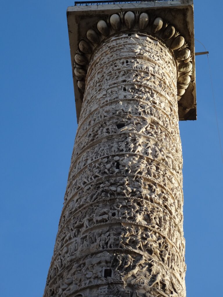 A photograph of a tall column which is inlayed with carvings of a parade of people and animals. The image was taken from below, panning up towards the top of the structure